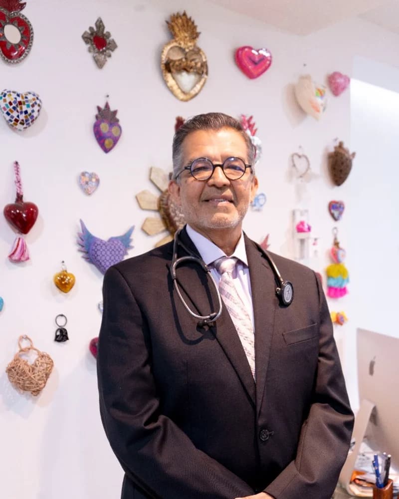 El Dr. Alfredo Estrada Suárez, con traje y estetoscopio, de pie frente a la pared de corazones de hojalata de su consultorio en Tuxtla Gutiérrez